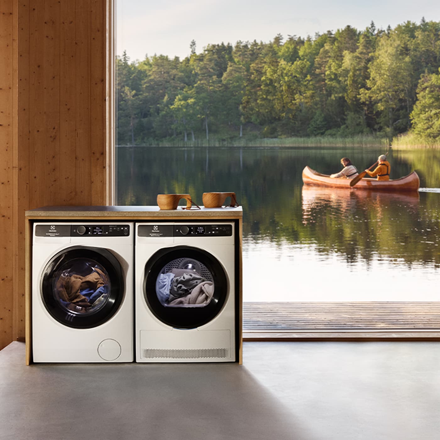 Pralka i suszarka obok siebie na tle dużego okna od podłogi po sufit przez które widać parę płynącą kajakiem